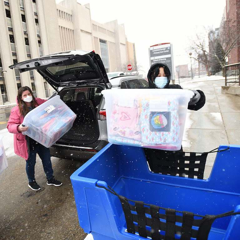 Two people transfer clear plastic bins with blankets and tote bags in them from the trunk of a car to a wheeled blue basket. 