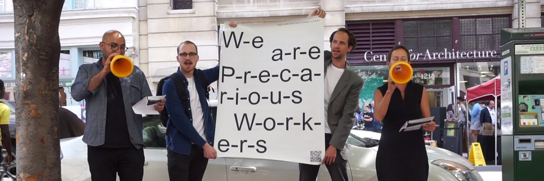 Two people holding a sign that reads: w-e-a-r-e-p-r-e-c-a-r-i-o-u-s-w-o-r-k-e-r-s, while two other people speak into microphones.