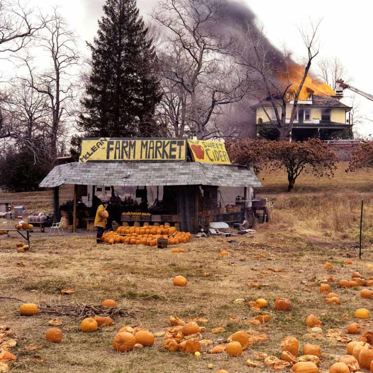 A farmer's market with pumpkin with a burning building in the background.