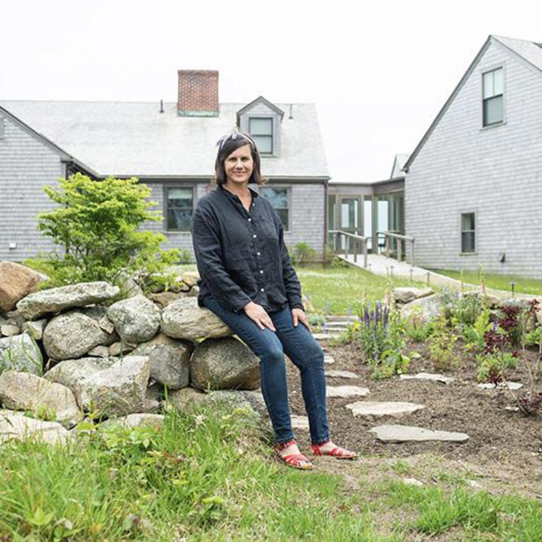 A person sits on a rock wall in front of two grey slatted buildings on Menemsha Beach. They are wearing a headband, black button up shirt, jeans, and red sandals. 