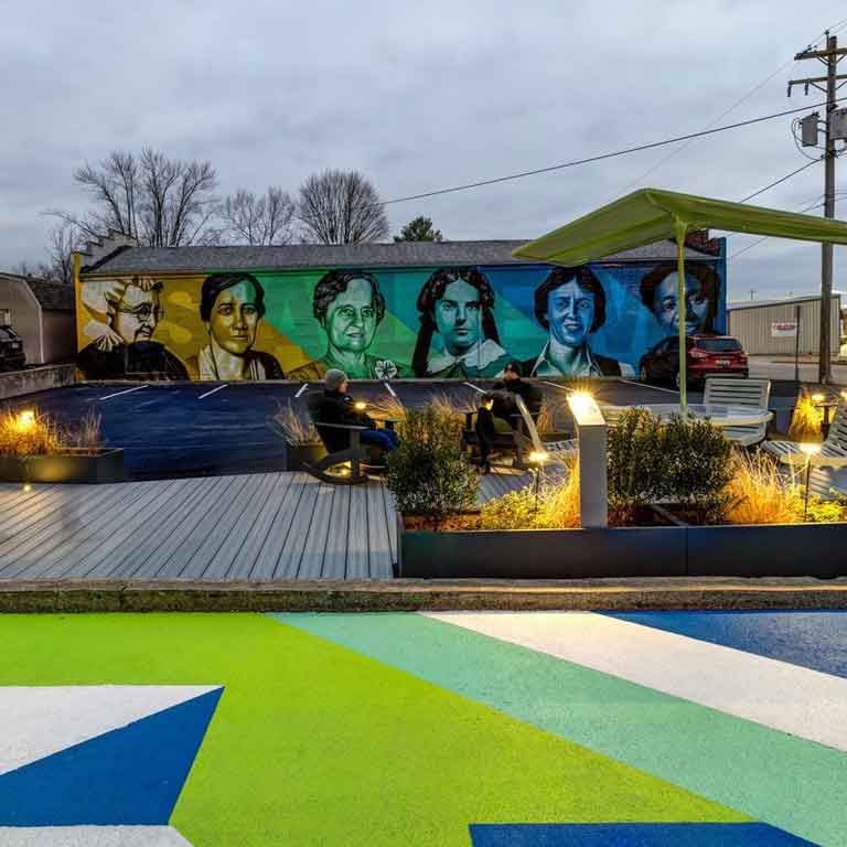 A mural of women in shades of blues and greens on a brick wall. The street is also painted in those colors, but in geometric shapes. There are covered areas to sit.