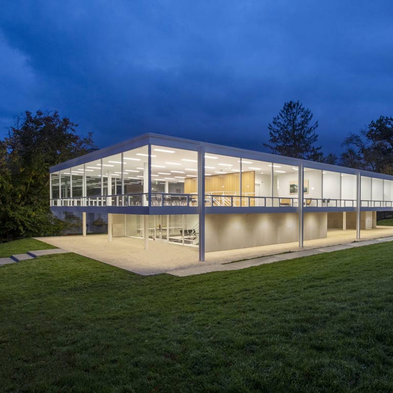 The Mies van der Rohe Building lit up at night. It is a white rectangular building against a deep blue sky. 