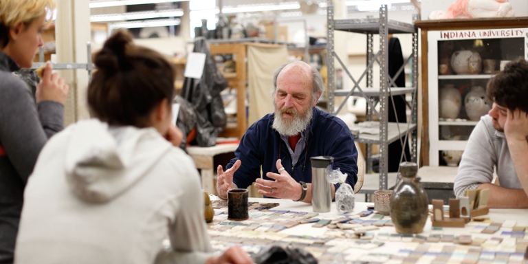 Students listen to a man talk at a desk in an art workshop.